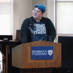 A person with blue hair standing at a podium, addressing an audience.